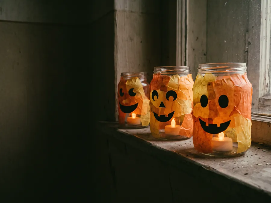 Drei Einmachgläser, außen mit orangem Transparentpapier beklebt, mit Gesichtern aus schwarzem Papier und LED-Teelichtern darin