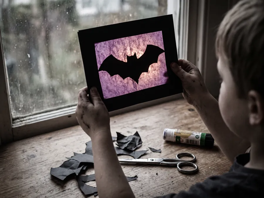 Kind hält ein Fensterbild gegen das Licht: schwarzer Kartonrahmen mit ausgeschnittener Fledermaus, von hinten mit violettem Transparentpapier beklebt