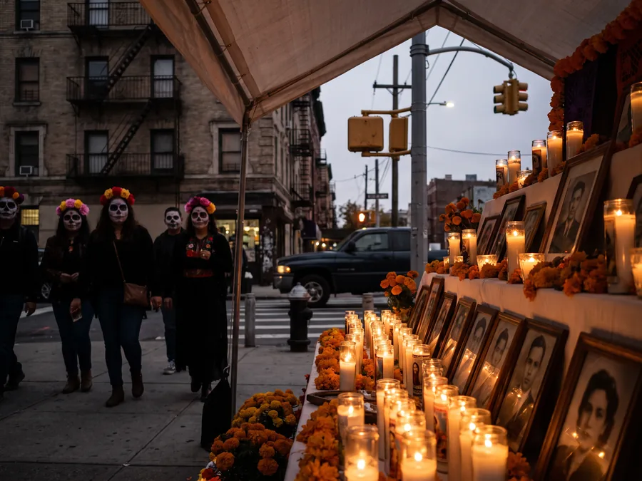 Langer Straßenaltar mit Kerzen in Gläsern, Ringelblumen und gerahmten Fotos auf einem Gehweg einer US-Stadt, dahinter Passanten mit Catrina-Schminke