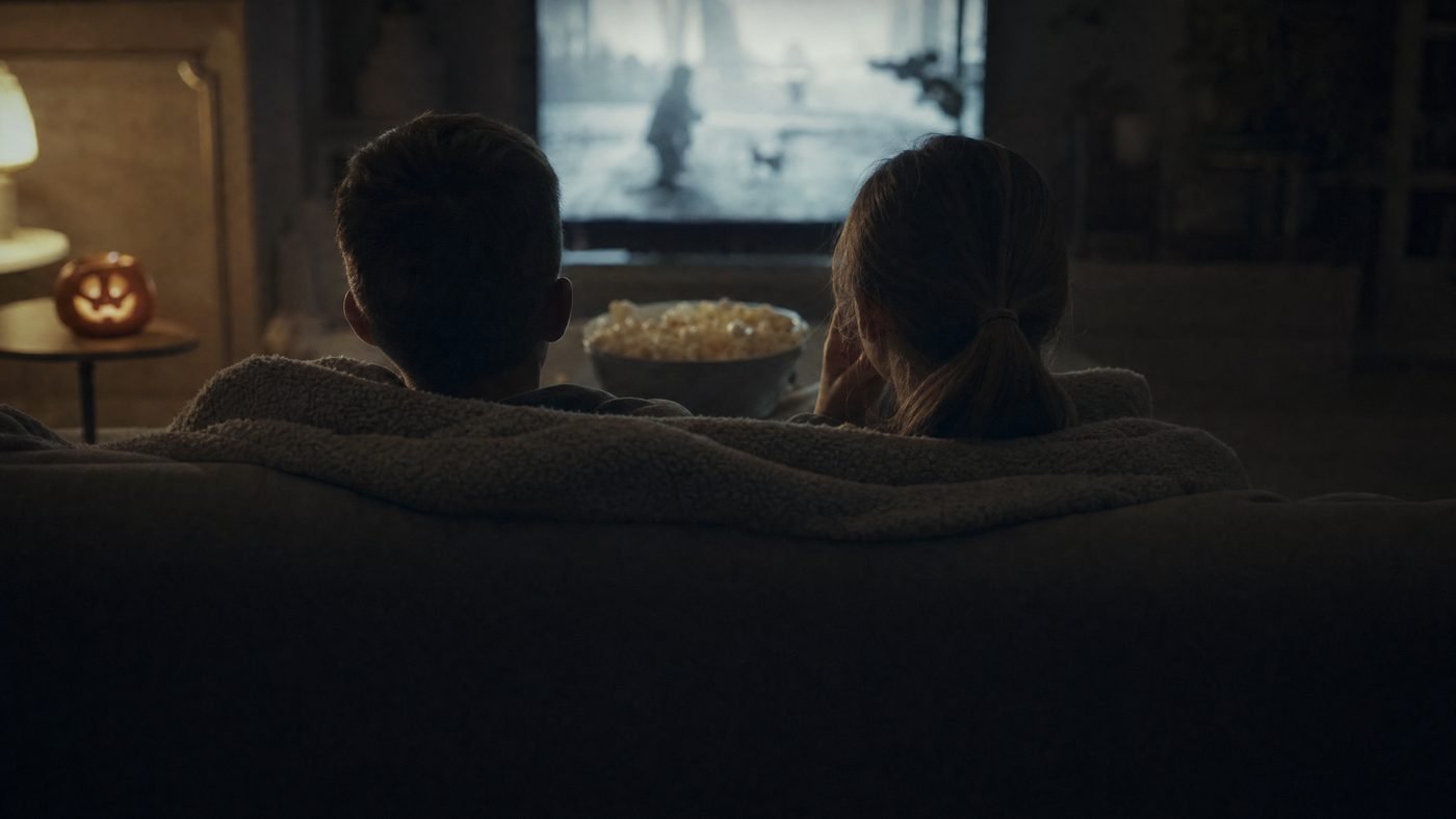 Zwei Kinder von hinten auf dem Sofa vor dem leuchtenden Fernseher, Popcorn und kleiner Kürbis daneben