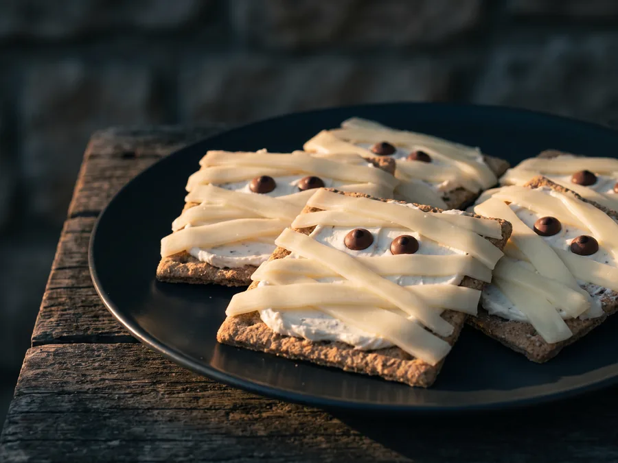 Cracker mit Frischkäse, Käsestreifen als Bandagen und zwei Zuckeraugen